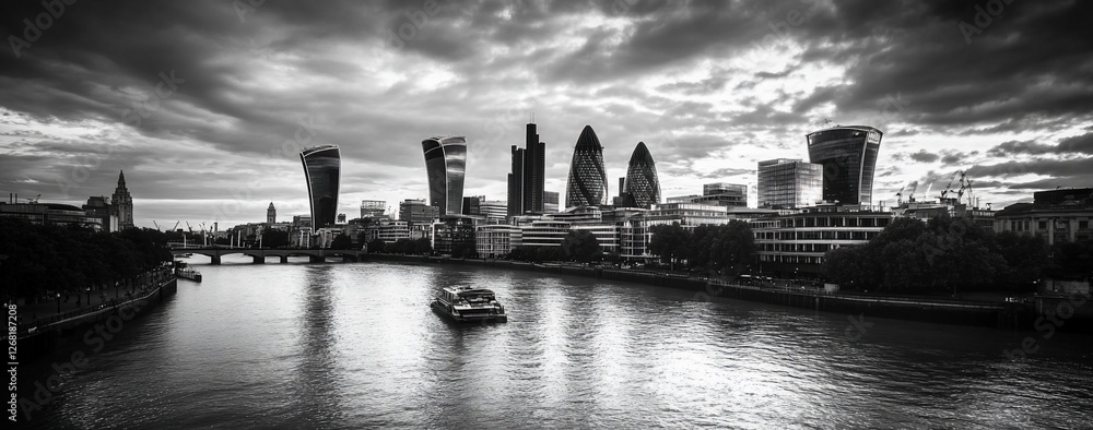 Wall mural Black and white panoramic view of London skyline at sunset, featuring cityscape, river, boat, and dramatic clouds.