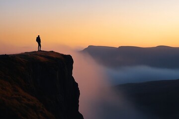 Silhouetted hiker on clifftop overlooking misty valley at sunrise.