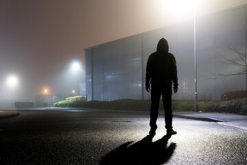 A sinister hooded figure, silhouetted by street lights on an empty industrial estate on a mysterious foggy night