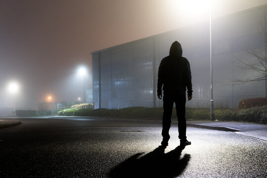 A sinister hooded figure, silhouetted by street lights on an empty industrial estate on a mysterious foggy night