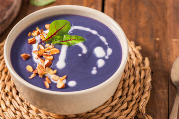Purple soup garnished with chopped nuts, a drizzle of cream, and a green leaf, served in a beige...