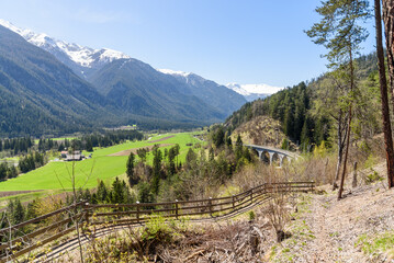 Narrow fenced path overlooking a large valley in the Swiss Alps on a sunny spring day. A stone railway viaduct is visible in distance.