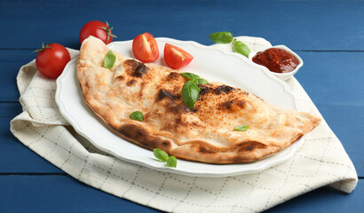 Tasty calzone with basil, tomatoes and sauce on blue wooden table, closeup