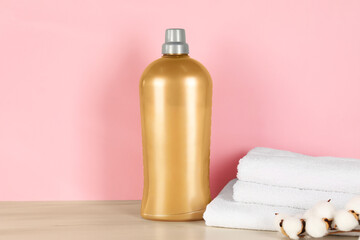 Bottle with laundry detergent, stack of clean towels and cotton flowers on wooden table against pink background