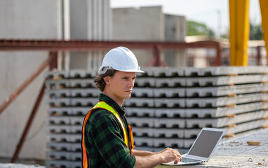 Fototapeta premium Civil engineer in safety suit is using his computer or laptop to check the quality of slabs and precast floorboard in production line. A male engineer with his computer using technology in a factory.