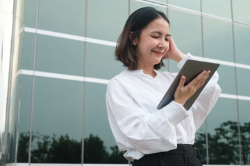 Happy woman in white shirt holding tablet, standing in front of modern glass building, reflecting clear sky, enjoying outdoor work environment.