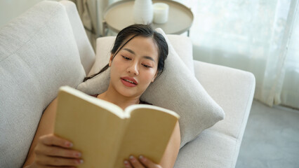 Carefree young woman relaxing on her couch while reading a book in a cozy, bright living room.