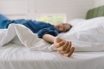 A woman lies comfortably on her back in bed, wearing a blue shirt, smiling as she stretches. Sunlight gently illuminates her peaceful expression, enhancing a serene morning atmosphere.