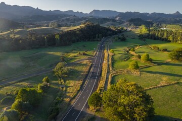 High-angle view of a rural road winding through lush green fields and forests, with mountains in...