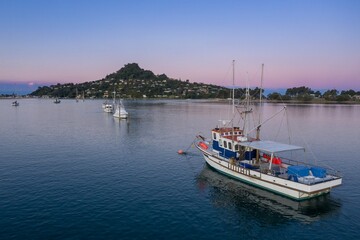 Fototapeta premium Tranquil morning scene on the water. Fishing boat navigates calm waters near coastal town. Peaceful harbor at dawn. Tairua, Coromandel Peninsula, New Zealand