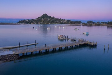 Wooden pier extends into calm water, boats moored nearby, tranquil coastal scene at dusk. Tairua,...