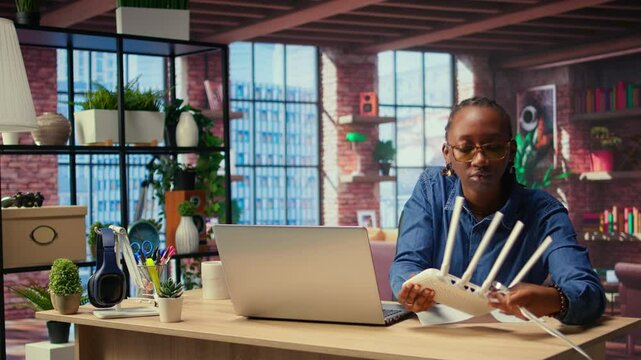 Black woman at home examines the modem and troubleshooting guide book, struggling to fix Wi Fi router and restore her internet connection for remote freelance tasks. Signal loss. Camera A.