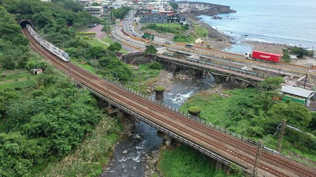 Aerial view of express of EMU 3000 and a cargo train dashing thru a bridge across a river by the mountainside and entering a tunnel with Daxi Fishing Harbor in background, in Toucheng , Yilan, Taiwan