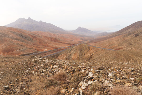 A barren, rocky mountain landscape in Fuerteventura under a cloudy sky. The mountains are brown and textured, with sparse vegetation in the foreground