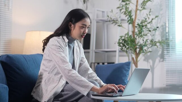 A woman is sitting on a couch and working on a laptop. She is wearing a white jacket and she is focused on her work. The room has a blue couch and a potted plant in the background
