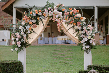 Wedding arches of orange, pink, white flowers, and cream fabrics. The wedding arch stands for a wedding ceremony held in the garden on the grass during the morning hours