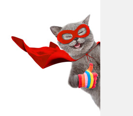 Happy cat wearing superhero costume looking above from behind blank white banner and showing thumbs up gesture. Isolated on white background © Ermolaev Alexandr
