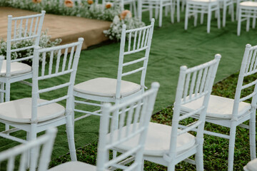 White chairs placed in a wedding setting in the garden and grass. Brown stage decorated with flowers