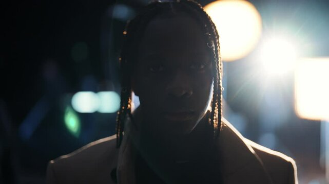 Portrait of black male fashion model on film set, mysterious man looks at camera . Stylish African American male person standing in darkness, gaffer adjusting soffits in background, contour light