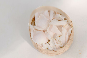 White and light pink rose petals are placed in a woven basket on a white background