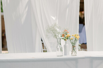 Fresh flowers in three vases are used for wedding decorations placed on a table covered with a white tablecloth. Additionally, there is a white cloth adorning the backdrop