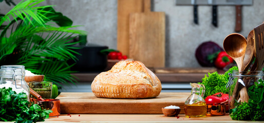 Rustic whole grain wheat sourdough bread on wooden cutting board.. Kitchen table with vegetables and herbs against the background of modern classic kitchen
