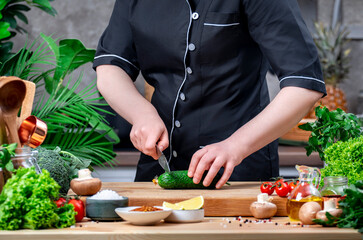 Chef cuts fresh cucumber with Japanese knife. Cozy kitchen with wooden table, kitchenware, vegetables, herbs and ingredients for cooking. Home food, cooking, recipes, food blogging