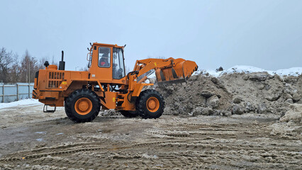 A heavy-wheeled excavator is loading collected dirty snow from the streets into a snow-melting machine