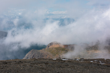 Misty aerial top view above abyss to sunlit big rocky hill top on green ridge spur in low clouds against large mountain range silhouette in cloudy blue sky. High mountains in foggy changeable weather.