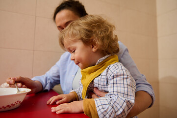 Mother Feeding Her Child at Home, Child Wearing Yellow Bib, Embracing Loving Interaction During...