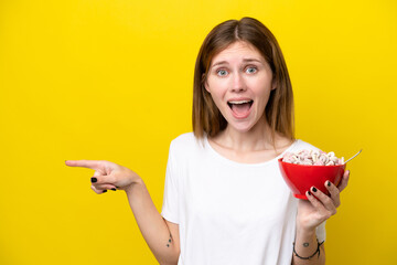 Young English woman holding cup of coffee isolated on yellow background surprised and pointing finger to the side