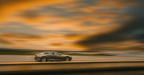 A brown sedan is driving on a highway at high speed. Side view of a moving car, blurred sunset background with orange clouds.