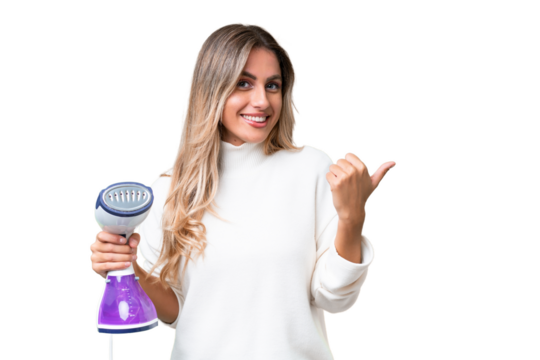 Young Uruguayan woman holding an iron over isolated background pointing to the side to present a product