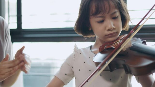 Asian young girl practices violin with focused dedication, guided by a supportive teacher showing music education, classical music, and the bond between student and mentor for nurturing talent.