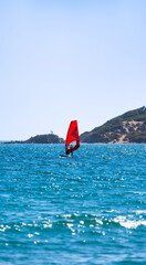 Windsurfer with red sail sailing in the sea with blue water and tourists sunbathing on the rocky coast in the background in Tarifa, Spain.