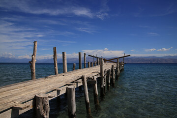 Fototapeta premium A weathered wooden pier extends into the crystal-clear blue sea, surrounded by mountains under a bright sky.