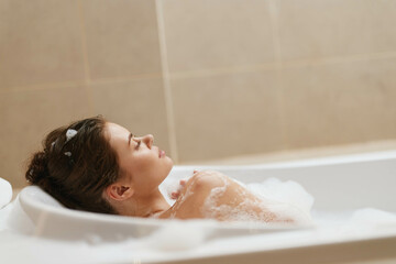 Young woman relaxing in a bubble bath, enjoying a moment of self-care and tranquility, with soft lighting creating a calming atmosphere in a modern bathroom.