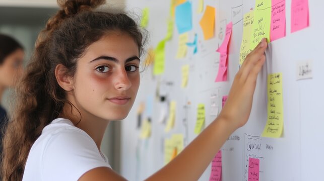 Focused Teenager Planning: A young woman with a focused expression adds a sticky note to a whiteboard covered in colorful notes, showcasing her dedication to planning and organization.
