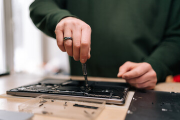 Cropped shot of electronics repair professional carefully disassembling laptop motherboard, precision screwdriver in hand, surrounded by various components on workshop table, close-up.