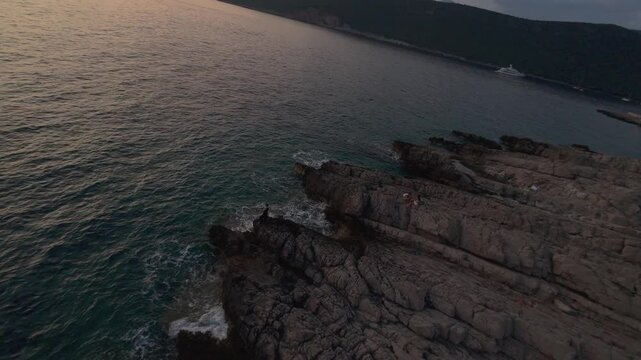 FPV drone circling around a woman in black standing on rocks by the sea with waves from the Lustica Bay near Fort Arza and Mamula Island at sunset, Montenegro, Europe