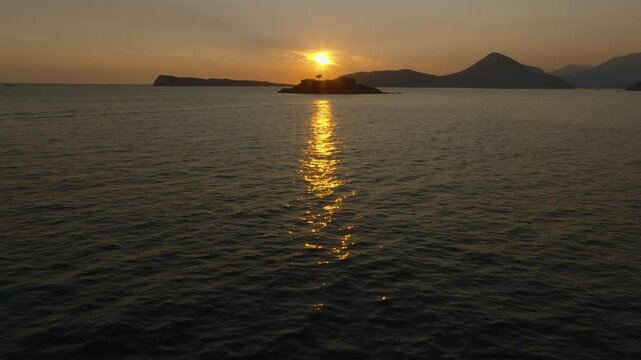 Cinematic drone view with camera rise from the reflection of the sunset on the water, leading to Mamula Island with silhouettes of mountains, Montenegro, Europe