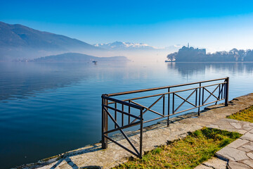 Ioannina Lake view with a metal railing in the foreground, leading to calm blue waters reflecting a misty landscape with distant mountains and Ioannina Island silhouetted on the left.