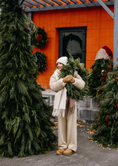 Joyful woman in a cozy teddy jacket, walking and holding a greenery wreath among evergreen trees.