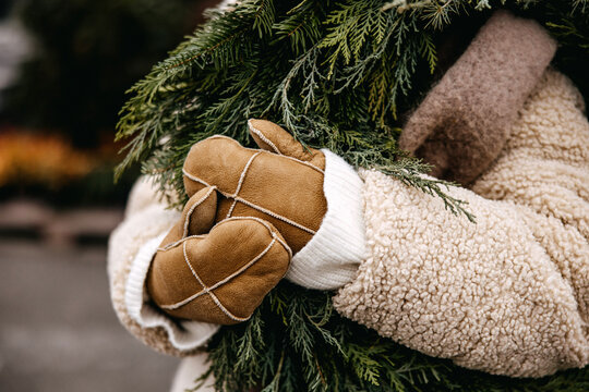 Close-up of cozy brown shearling gloves and a fluffy beige coat holding a lush green holiday wreath