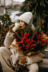 young woman in cozy neutral layers, holding a festive Christmas bouquet and a cup of coffee