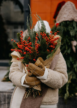 Woman in a cozy winter outfit with mittens holding a bouquet of red berries and greenery, festive holiday vibe.