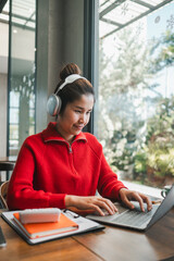 Smiling woman wearing headphones, working on a laptop in a bright, modern cafe with large windows and greenery outside.