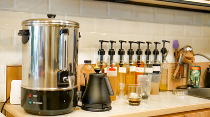 The kitchen of a cafe has a hot water dispenser, a teapot, and a collection of syrup bottles to flavor the drinks.