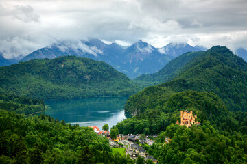 view of a castle in the mountains and a lake in bavaria germany. A mountain range with a lake in the middle. The lake is surrounded by trees. The mountains are covered in snow