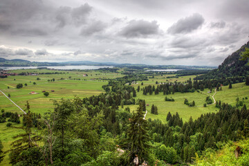 A large field with trees and a lake in the background in Bavaria Germany. The sky is cloudy. There are a few houses in the distance